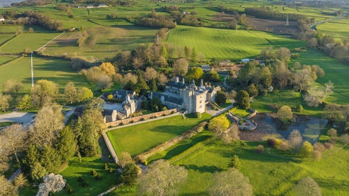 View of Sizergh's grey stone manor house, outbuildings, garden and surrounding estate from above. It's a sunny spring day, and the grass is green.